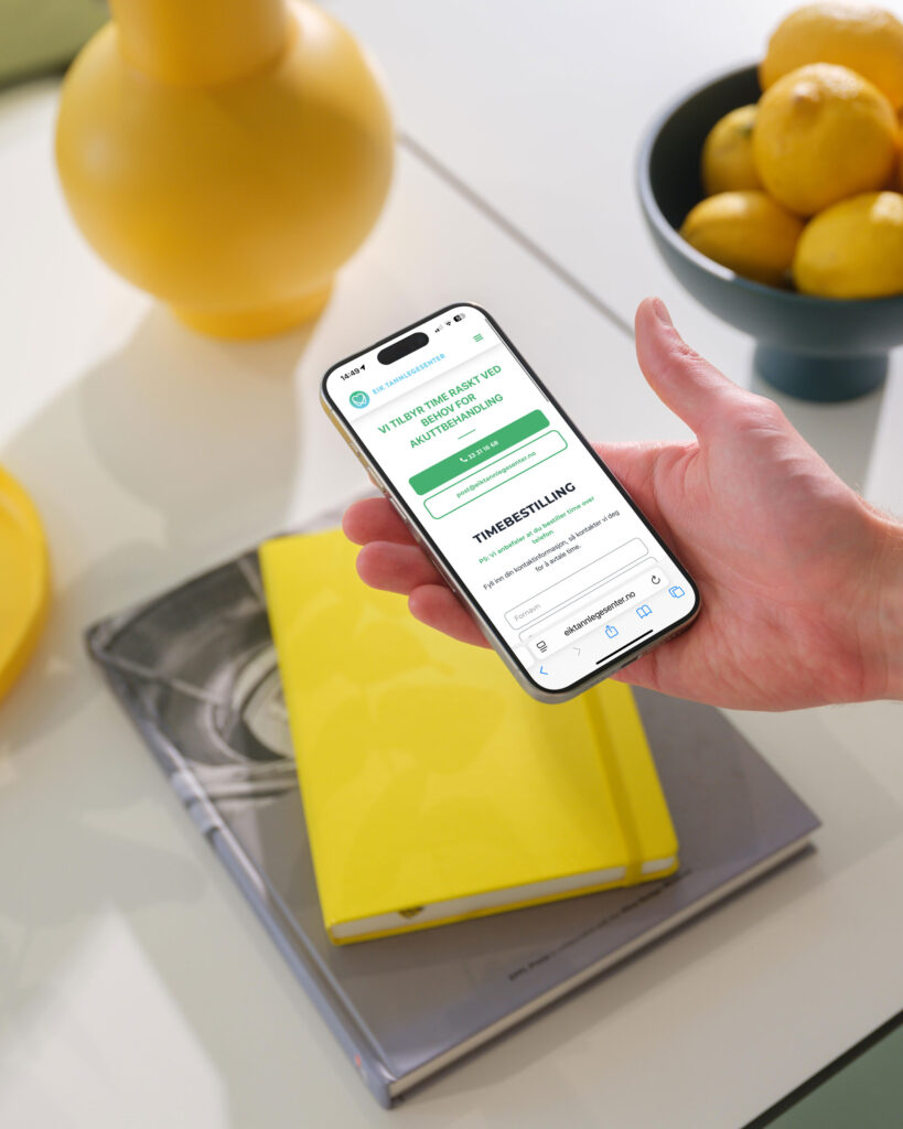 Bilde av en hånd som holder en iphone som viser hjemmesiden til eik tannlegesenter foran en gul skriveblokk